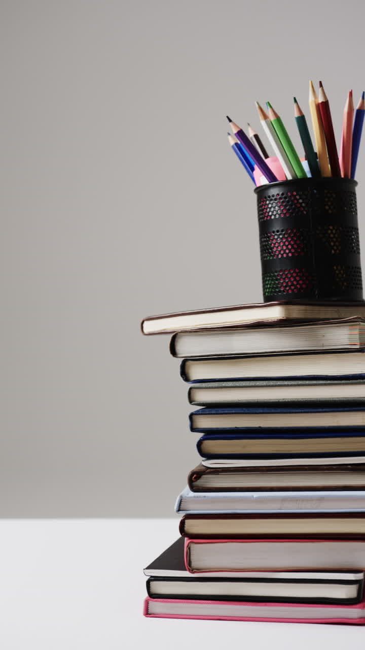 Vertical video of cup with crayons on stack of books on grey background, in slow motion