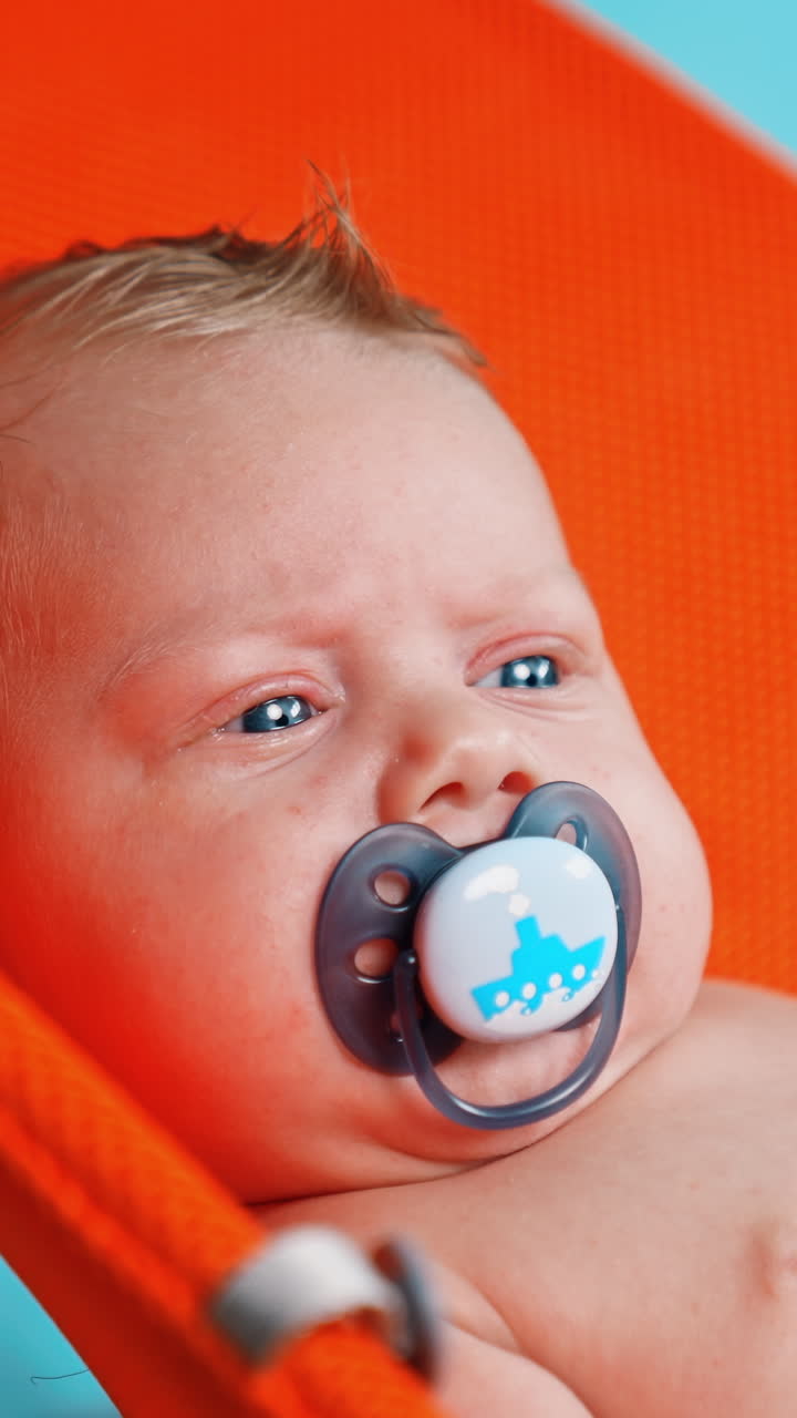 Unhappy blue-eyed blond Caucasian infant with a pacifier in mouth. Portrait of a kid in orange chair. Close up. Vertical video