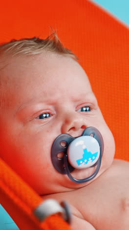 unhappy blue-eyed blond caucasian infant with a pacifier in mouth. portrait of a kid in orange chair. close up. vertical video