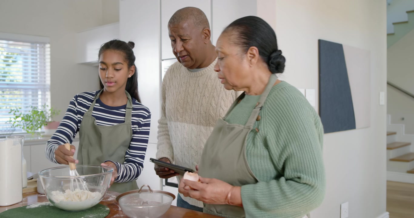 African American seniors guiding teen mixing batter at kitchen island, checking tablet, adding milk