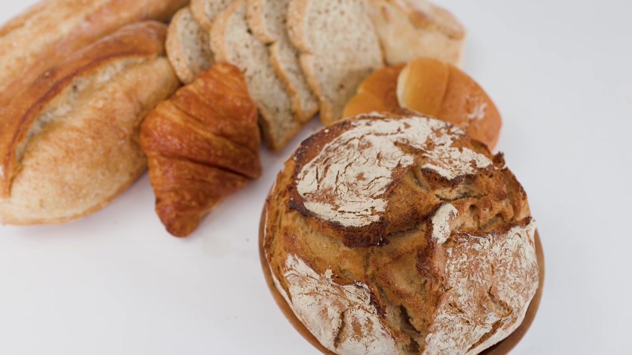 Lenta rotazione della telecamera rivela una pagnotta di pane a lievitazione naturale, un croissant e una baguette su una superficie bianca pulita.