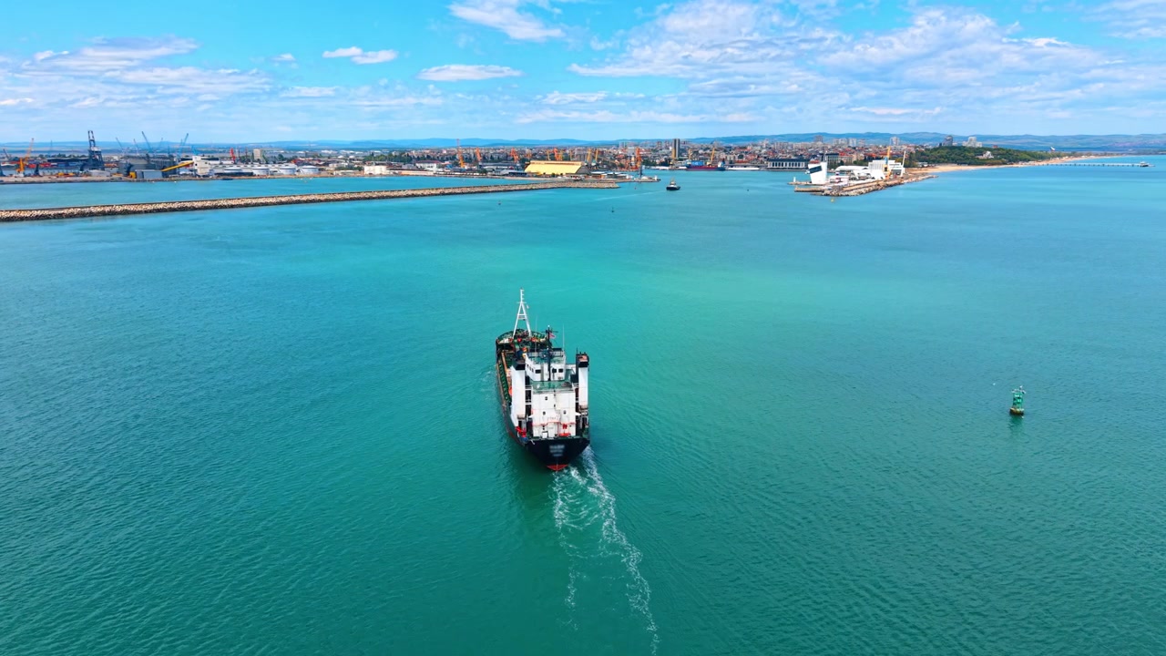Cargo vessel sailing through sea channel into port aerial view. High angle drone shot of cargo ship entering harbor channel with city port and long breakwater in background.