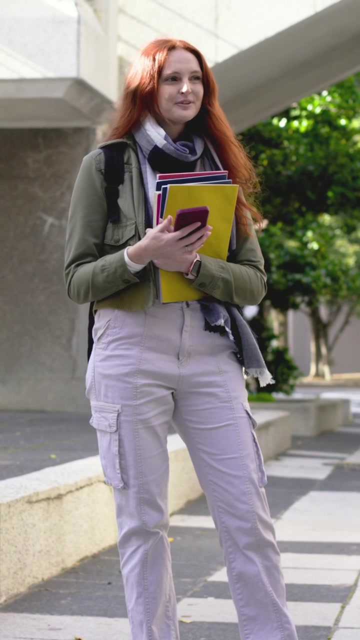 Video verticale: Donna in giacca che cammina nel campus, legge un messaggio sul telefono, con una cartella gialla, in attesa.