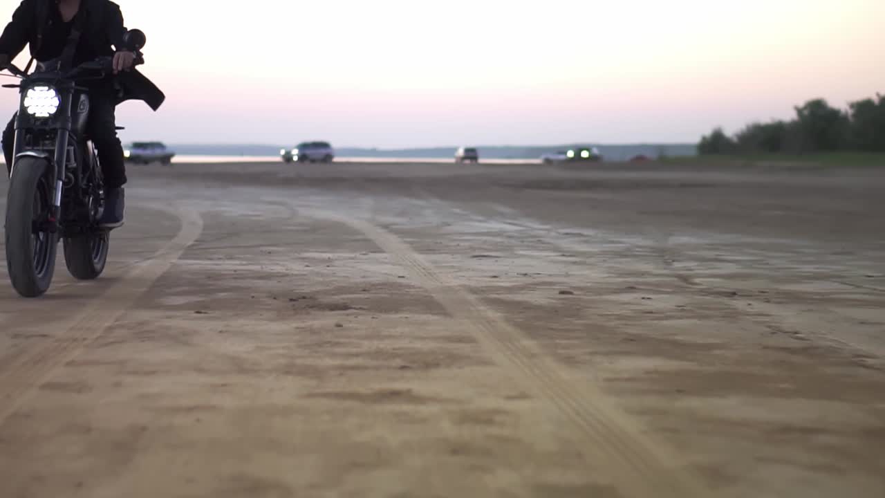 Motorcyklist med hjelm og jakke, en mand kører på en sportsmotorcykel om aftenen i lyserød skumring med nogle biler i baggrunden.