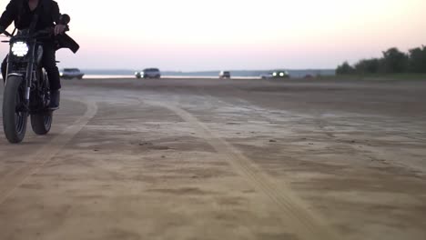motociclista en casco y chaqueta, el hombre está montando en motocicleta deportiva por la noche, crepúsculo rosa con algunos coches en el fondo