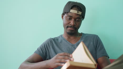 Close up of student in casual shirt and cap sitting on red couch with crossed leg, opening novel to read, head slightly tilted downward in quiet indoor setting with turquoise wall in background