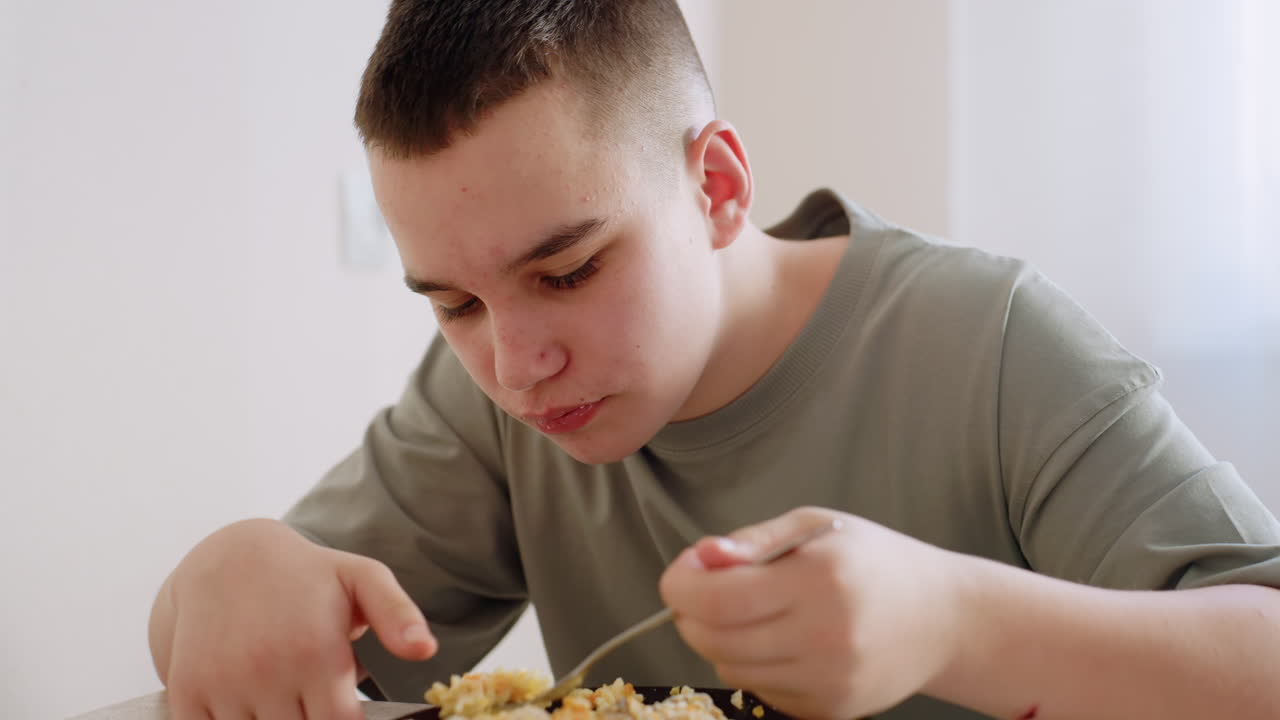 Primo piano di un bambino che mangia uova strapazzate con la forchetta al tavolo da pranzo, espressione concentrata mentre mastica il cibo, luce naturale che filtra attraverso la tenda e illumina il semplice stile di vita domestico