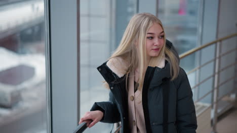 woman in jacket looking contemplative with soft gaze near glass building, exuding quiet confidence, her long blonde hair flows over her shoulders as she appears deep in though