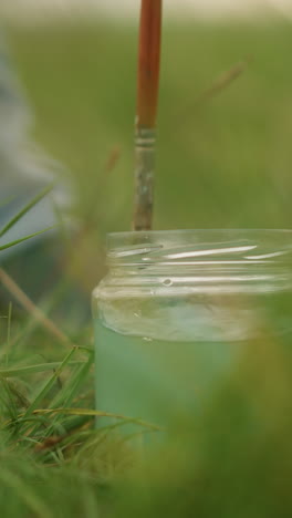 una toma de primer plano de la mano de un artista sumergiendo un pincel en un frasco de agua, en el fondo de la hierba verde exuberante. la imagen captura el momento de enjuagar el pincel