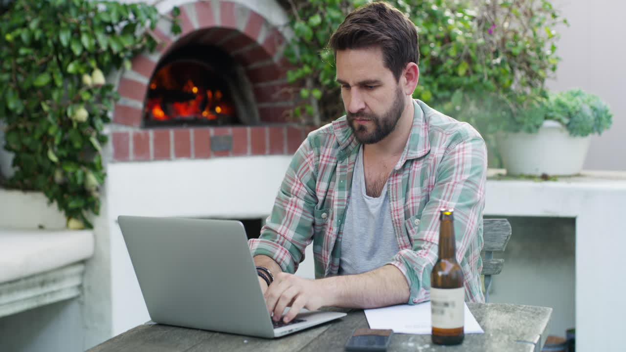 hombre trabajando en una computadora portátil en un patio con horno de pizza