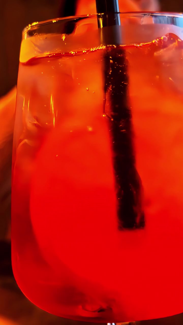 Primo piano estremo di un vivace cocktail Aperol Spritz arancione con cubetti di ghiaccio, fetta d'arancia e cannuccia nera in un bicchiere da vino sotto una calda e suggestiva illuminazione da bar.