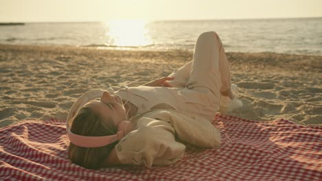 una chica rubia con ropa ligera y auriculares rosados escucha música y se acuesta en un paquete en una playa soleada por la mañana