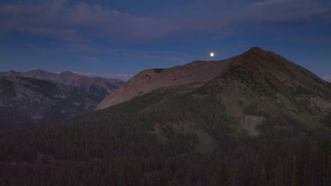 a full moon sits just above a mountain peak in the colorado rockies on a beautiful night