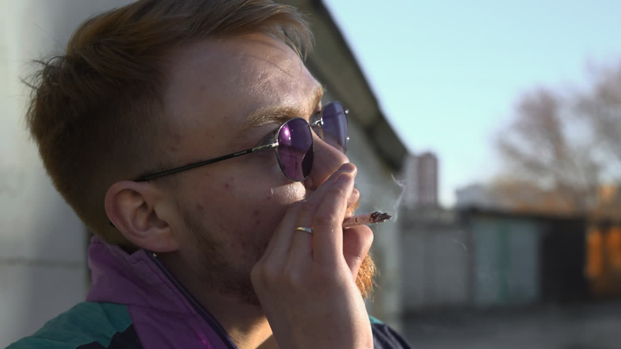 Jonge man rookt op het dak bij zonsondergang terwijl hij in de verte staart, roodharig bebaard profiel met ronde zonnebril.
