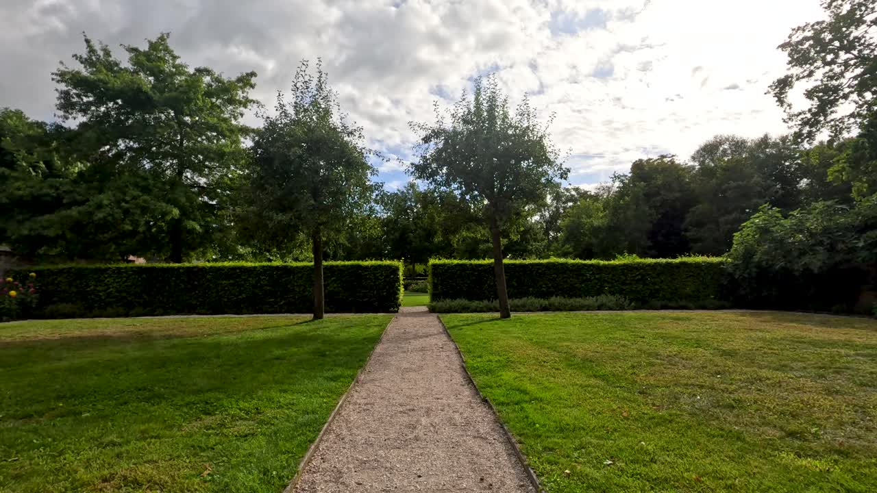 A steady forward camera movement along a gravel path in a formal botanical garden, with symmetrical greenery and soft daylight under partly cloudy skies