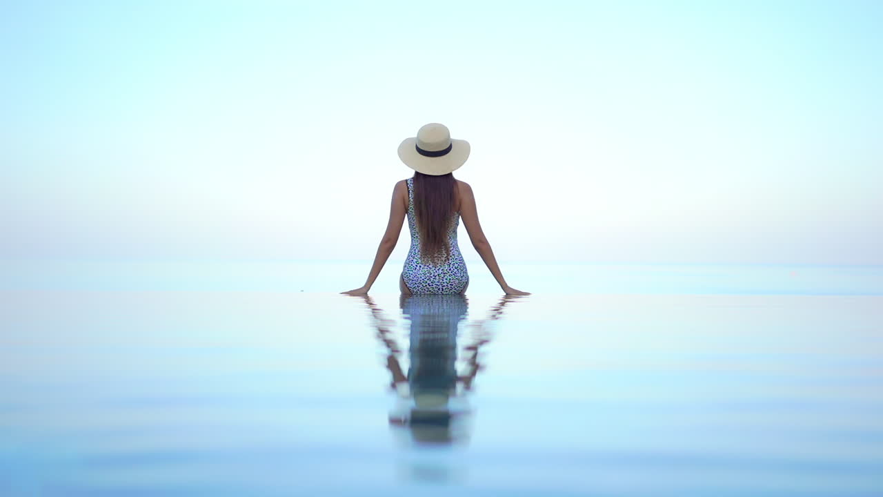 카메라를 등지고 있는 매우 건강하고 섹시한 여성이 수평선을 바라보며 수영장 가장자리에 앉아 있습니다.