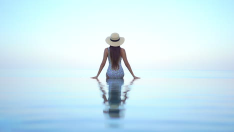 A-very-fit-and-sexy-woman-with-her-back-to-the-camera-sits-on-the-edge-of-a-swimming-pool-as-she-looks-out-at-the-horizon