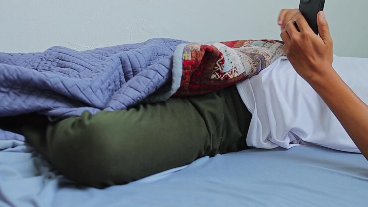 Persona che riposa sotto una coperta su un letto a pavimento si allunga per prendere lo smartphone.