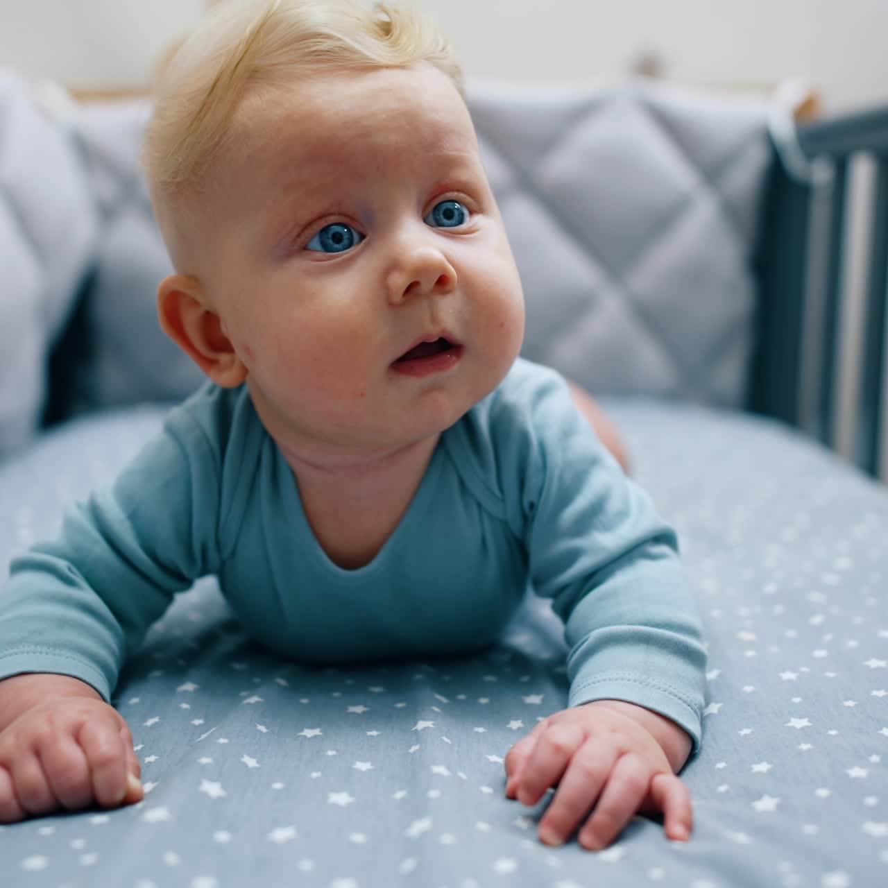 Blond babygutt i blå klær ligger på magen og støtter seg på hendene. Det søte barnet ser opp med overraskelse.