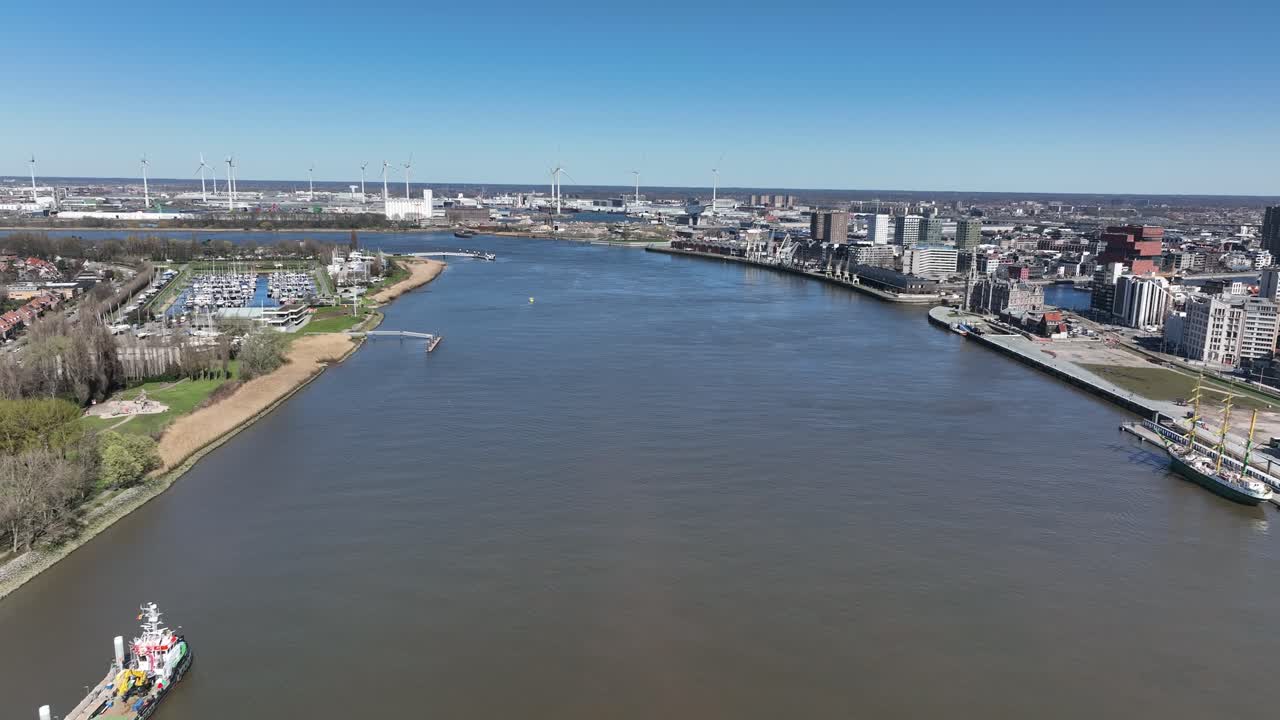 Aerial zoom in over Antwerp riverside showing marina, residential areas, and harbor with modern city buildings under clear blue sky