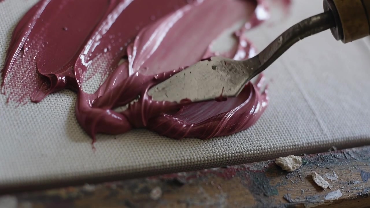 Pulling palette knife from right edge, artist shaping mauve paint on studio canvas