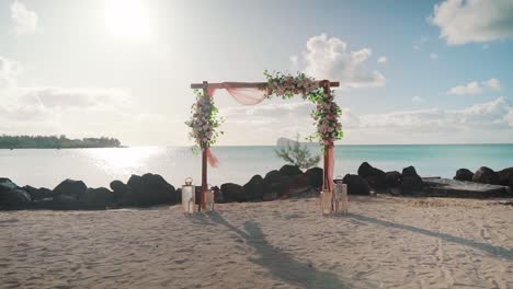un arco cubierto de flores para un evento de boda en una playa de arena y el océano al sol