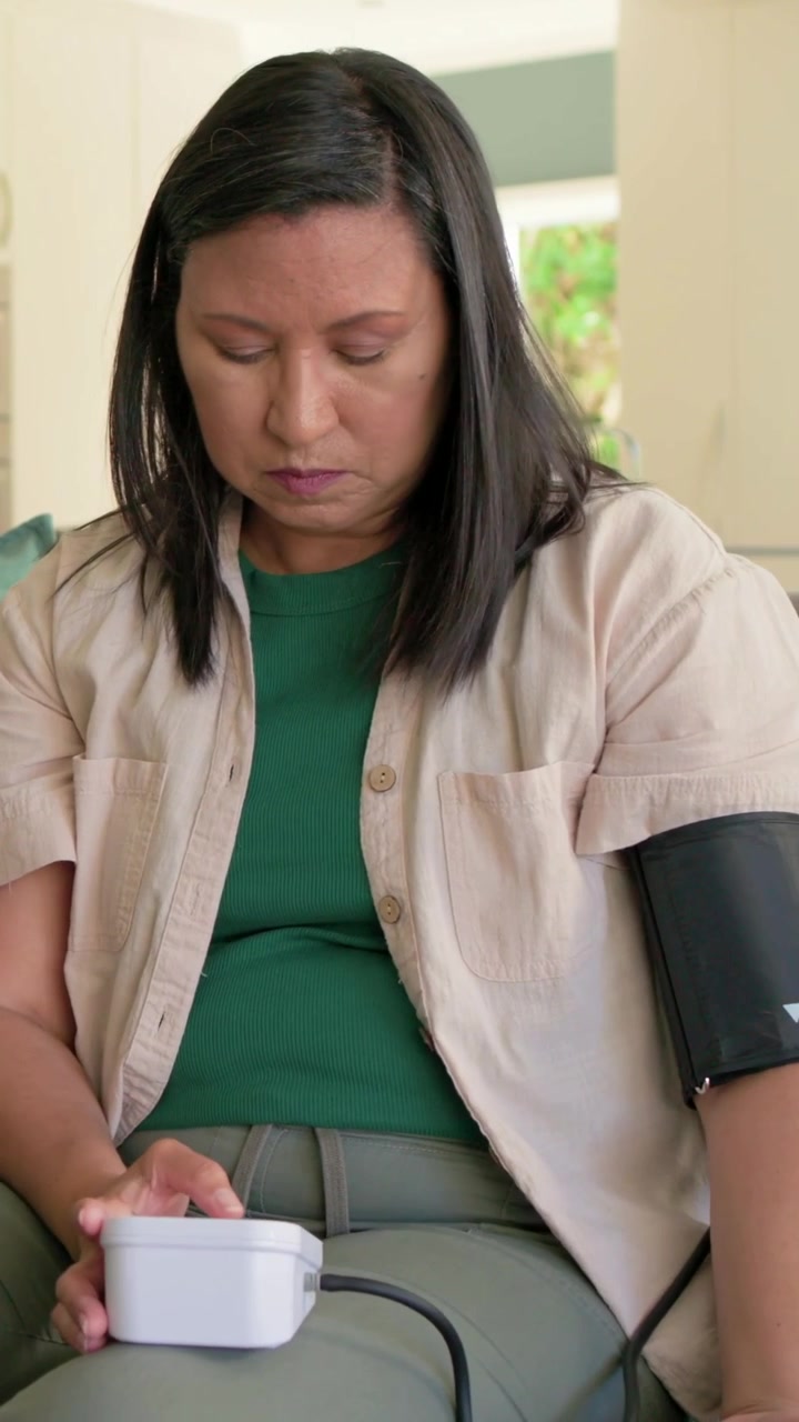 Video verticale: Una donna con una maglia verde si allunga, posiziona il bracciale sul divano, collega il monitor e misura la pressione sanguigna.