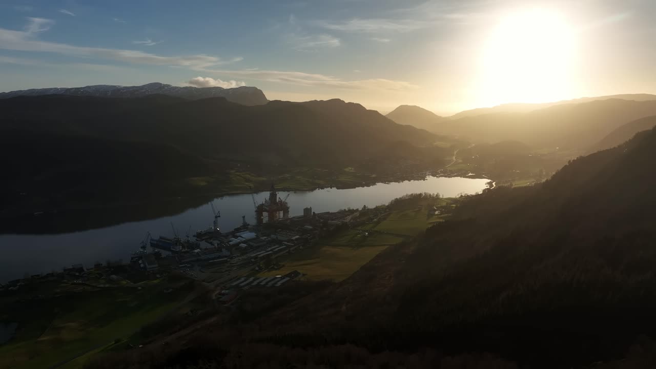 Aerial moving along mountainside toward Olensvag, Norway, revealing Westcon Yards, cranes, ships, and a drilling rig. Vibrant sunset over fjord and landscape