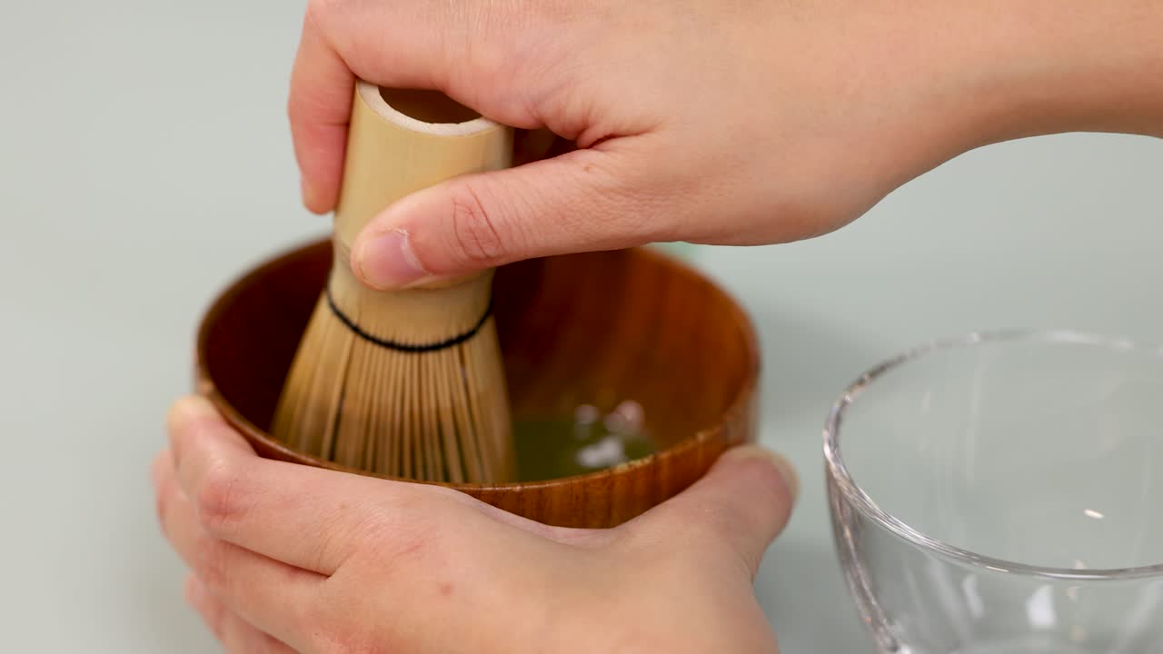 Hand whisking matcha green tea with bamboo whisk in wooden bowl, soft natural lighting, close-up