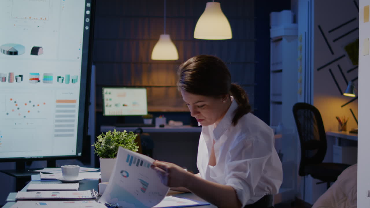 Workaholic businesswoman looking at reports presentation on monitor comparing with financial chart