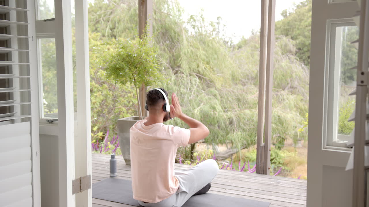 Meditating on yoga mat, man wearing headphones and relaxing on porch