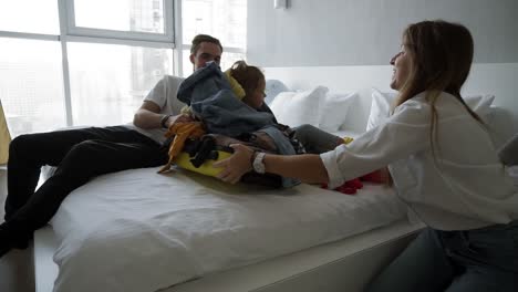 joven familia divirtiéndose mientras empaca para las vacaciones. hombre caucásico, mujer y niño en el dormitorio. niño pequeño está sentado en el pequeño