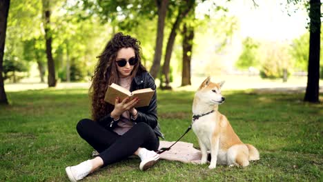 attractive young woman is reading book sitting on grass in park while her well-bred dog is sitting near her owner and looking around. leisure and animals concept.