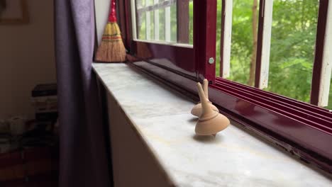 a woman rotate spin the wooden handmade spinning top at the red metal frame window in forest house country side village rural life in iran natural landscape kid game play craft day time cloudy weather