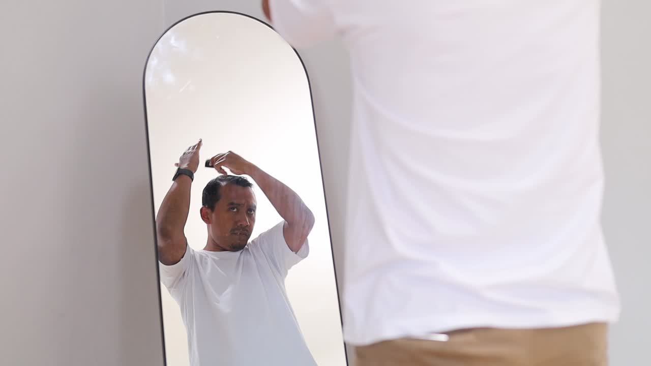 Uomo asiatico affascinante e sicuro di sé che si spazzola i capelli tenendo un pettine e guardandosi allo specchio in camera, concetto di cura e styling dei capelli.