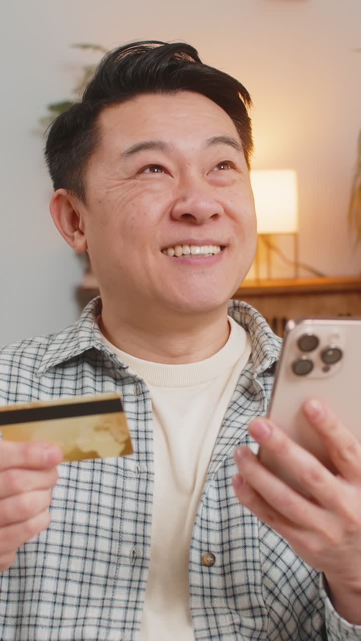 快乐的中年男子坐在沙发上使用信用卡和智能手机在线购物