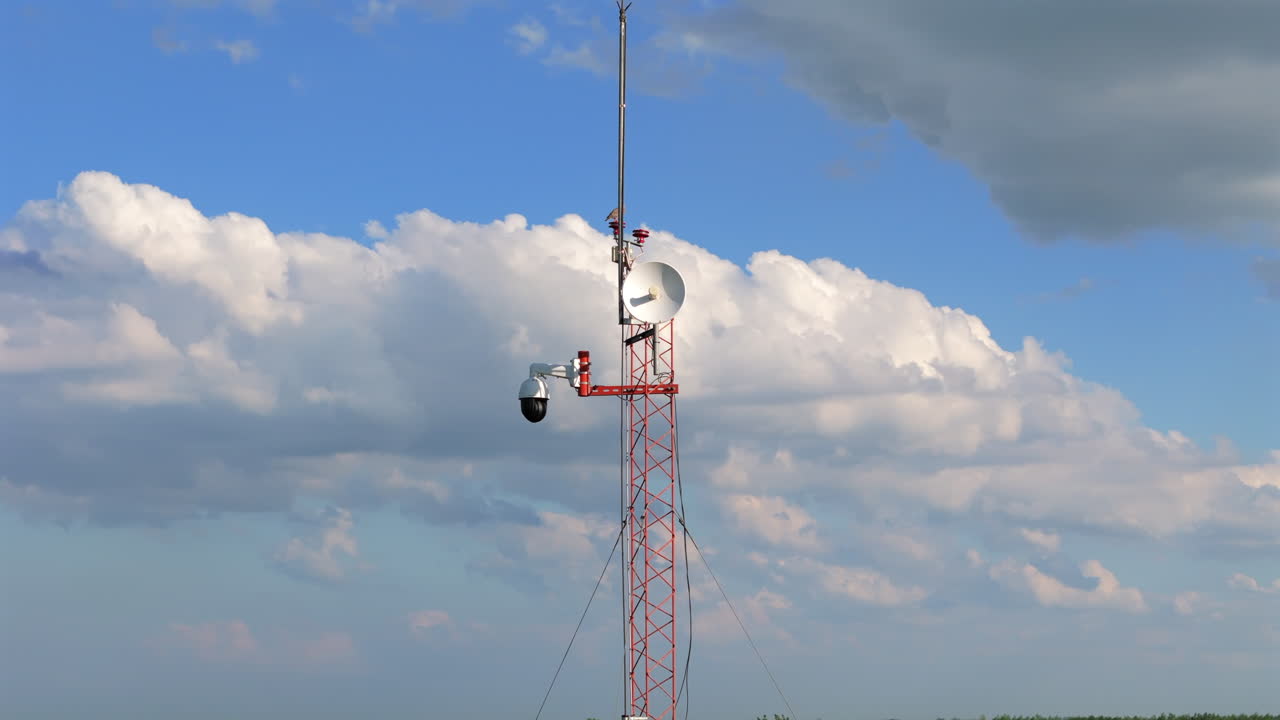 A drone orbits around a tall communication tower, showcasing its intricate structure and equipment like large satellite dishes and antennas.