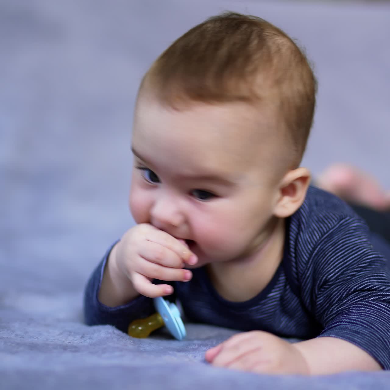 Tiny cute boy lies on bed and chews his pacifier. Little kid drops the nipple and starts to chew his fingers. Close up