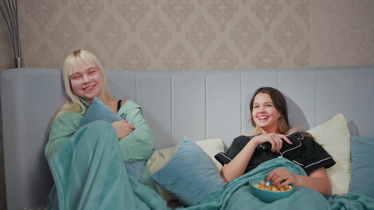 Two teenage girls in pajamas lying in bed, sharing laugh while watching TV, one holds remote control, other hugs pillow, popcorn bowl and plate with sandwiches rest on blanket in cozy bedroom setting