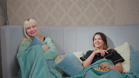two teenage girls in pajamas lying in bed, sharing laugh while watching tv, one holds remote control, other hugs pillow, popcorn bowl and plate with sandwiches rest on blanket in cozy bedroom setting