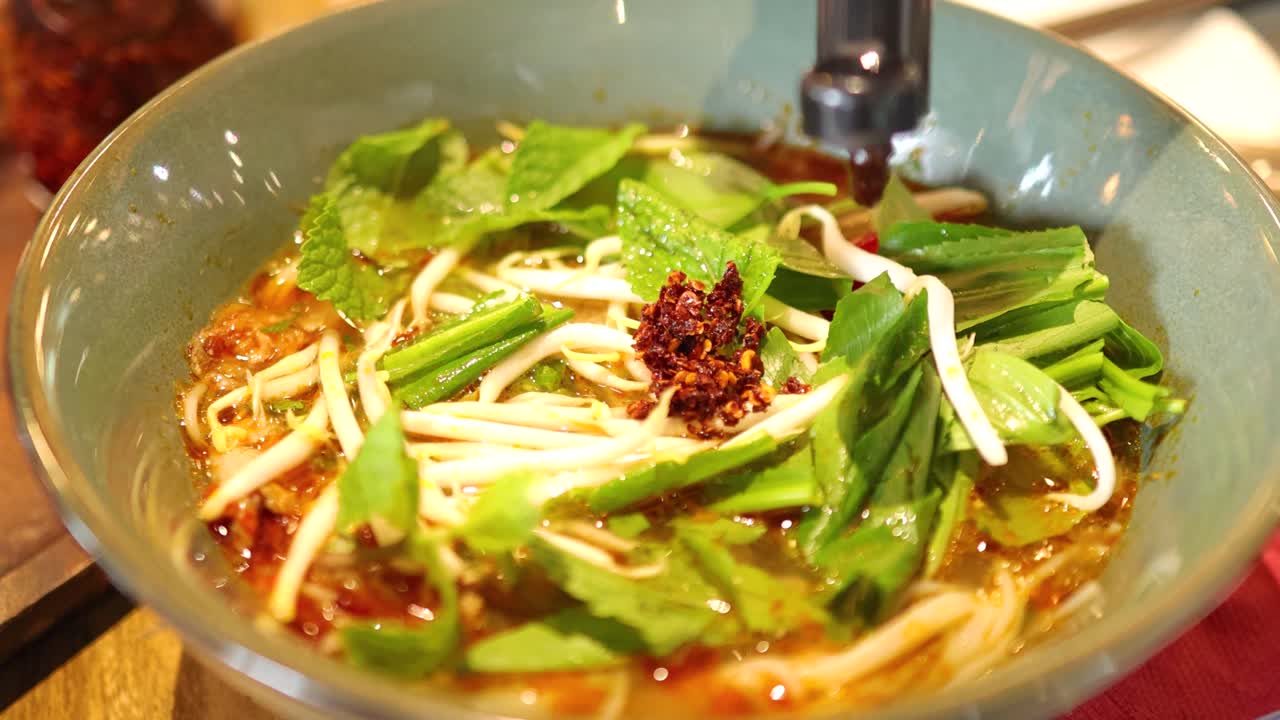A bowl of Vietnamese noodles is garnished with sauce and vegetables in a brightly lit restaurant setting