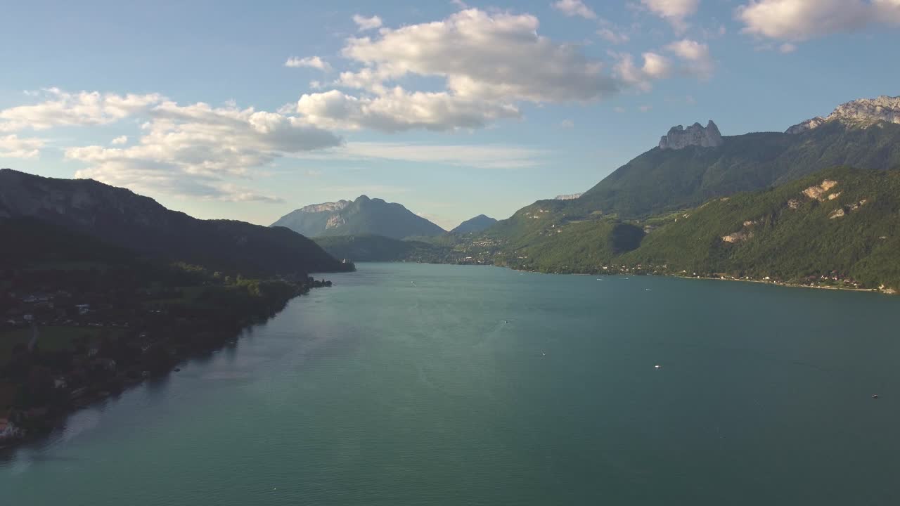 High view of lake Annecy with the sun shining on the surrounding mountains. Some clouds in there sky. Drone footage moves sideways to the left throughout clip