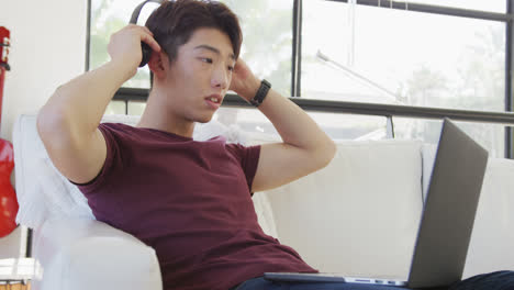 asian male teenager wearing headphones and using laptop in living room