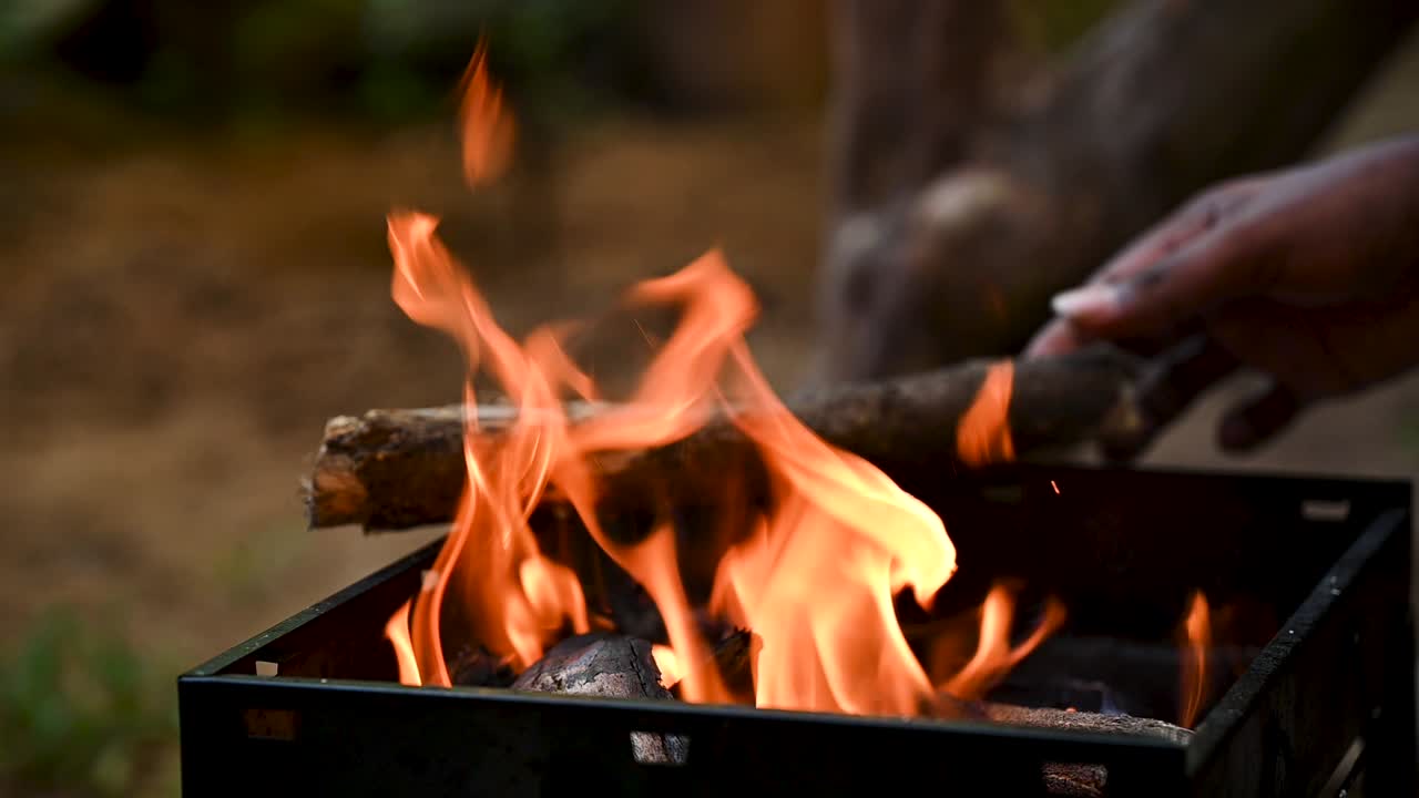 Slow-motion shot of placing firewood on a barbecue grill. Flames rise, creating a rustic fire for outdoor cooking. Perfect for barbecue, camping, lifestyle, food, and eco-friendly cooking visuals