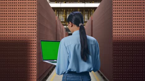 back view of asian female with red brick packed in stacks are stored, typing on laptop's keyboard with green screen and look at factory