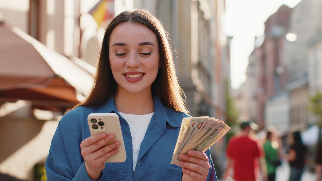 Happy young woman counting money dollar cash use smartphone calculator app in urban city street
