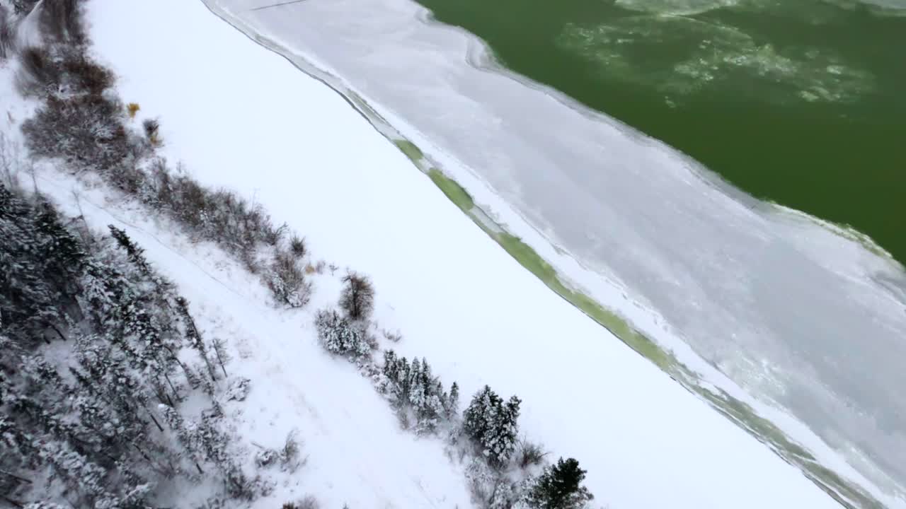 A breathtaking FPV shot performing a diving barrel roll, revealing snow-covered pine trees, a frozen river, and a man riding a snowmobile. Stunning winter wonderland captured in dizzying motion.
