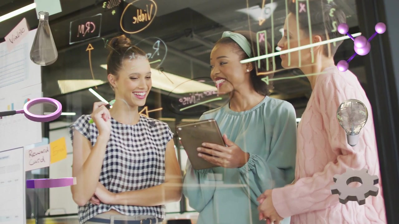 Diverse female team standing at glass drawing notes in office, teenage woman sharing tablet, marker