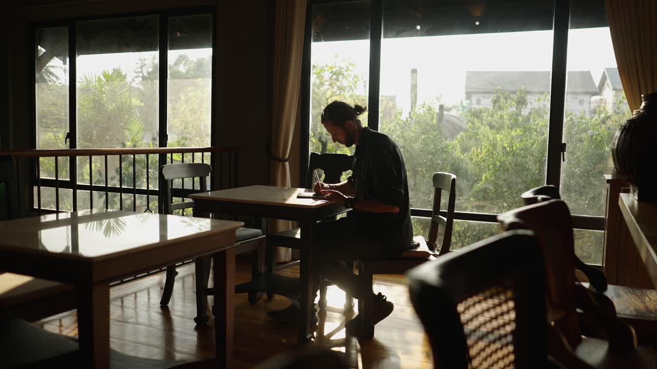 Man Writing in a Notebook at a Cafe by a Window