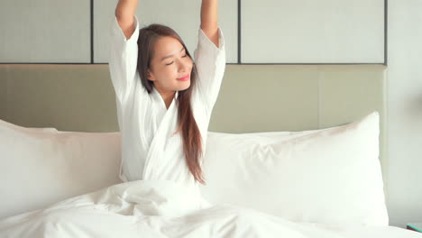 satisfied and smiling woman in bathrobe stretches sitting on bed